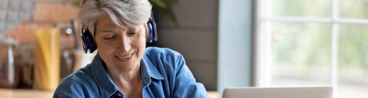 Lächelnde Frau mittleren Alters sitzt mit Kopfhörern vor dem Laptop und arbeitet.